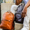 Two leather laptop backpacks, one brown and one black, placed on a person's lap who is seated on steps.