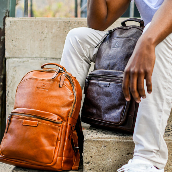 Two leather laptop backpacks, one brown and one black, placed on a person's lap who is seated on steps.